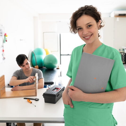 Portrait of beautiful physical therapist looking at camera smiling very happy while holding a tablet and patient at the background doing his therapy